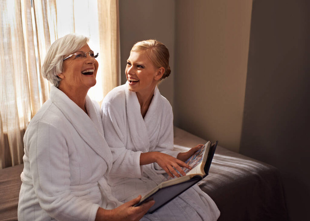 Grandmother, woman and laughing at photo album, home and happy for bonding and looking at memories. Senior lady, daughter and moment for self care, robe and bedroom for comfort and hospitality. - 写真・画像
