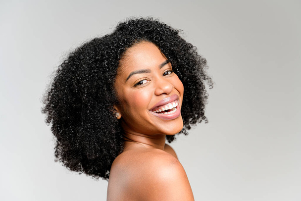 A profile view of an African-American woman with a beaming smile and lush curly hair, she exudes happiness and positivity against a muted backdrop, capturing a moment of pure joy. - Photo, image