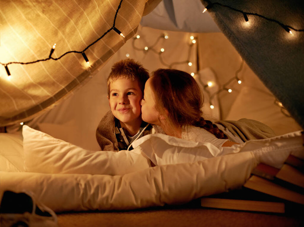 Children, happy and playing in tent at night with love, kiss and bonding for holiday adventure or vacation. Young boy, girl or kids by fairy lights, pillows and blanket at home with thinking or ideas. - Photo, Image