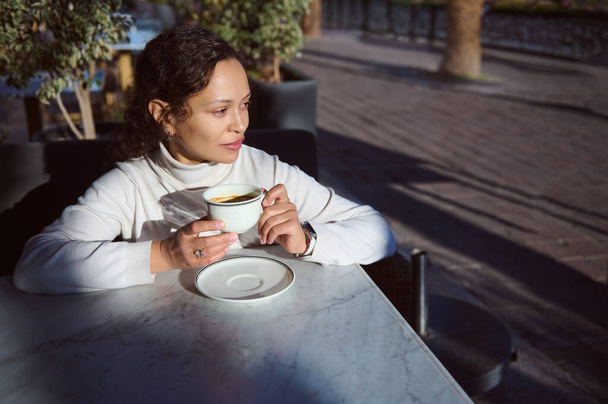 Многонациональная молодая стильная женщина пьет кофе на открытом воздухе в кафе, мечтательно глядя в сторону. Начните день с чашки бодрящего кофейного напитка. Людей. Стиль жизни. Деятельность на выходные - Фото, изображение