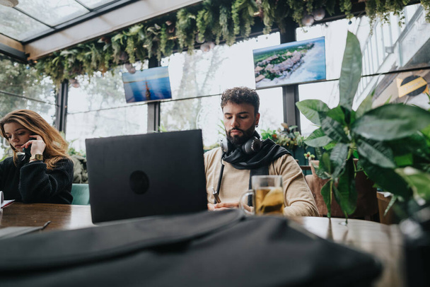 Ukierunkowani młodzi profesjonaliści zaangażowani w pracę w laptopach w przytulnej, wypełnionej roślinami miejskiej kawiarni. Koncepcja współpracy i przedsiębiorczości. - Zdjęcie, obraz