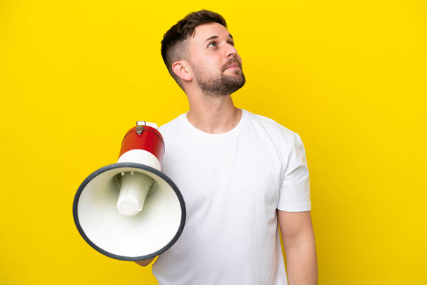 Young caucasian man isolated on yellow background holding a megaphone and looking up while smiling - Photo, Image