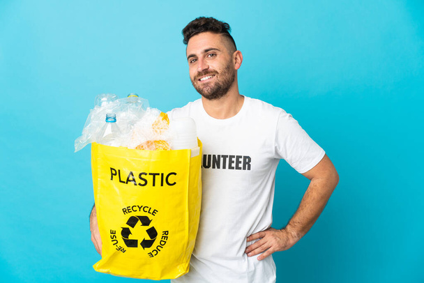 Hombre caucásico sosteniendo una bolsa llena de botellas de plástico para reciclar aislado sobre fondo azul posando con los brazos en la cadera y sonriendo - Foto, imagen