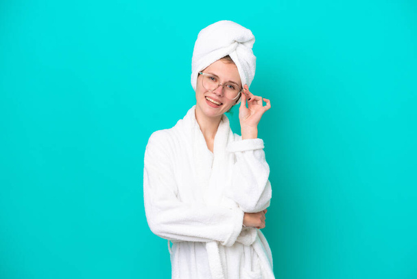 Young woman in a bathrobe isolated on blue background with glasses and happy - Photo, Image