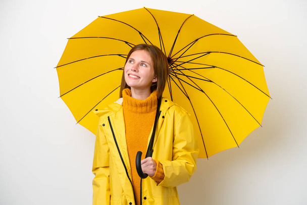 Yağmura dayanıklı ceketi ve şemsiyesi olan genç bir İngiliz kadın beyaz arka planda izole edilmiş bir şekilde yukarı bakarken bir fikir düşünüyor. - Fotoğraf, Görsel