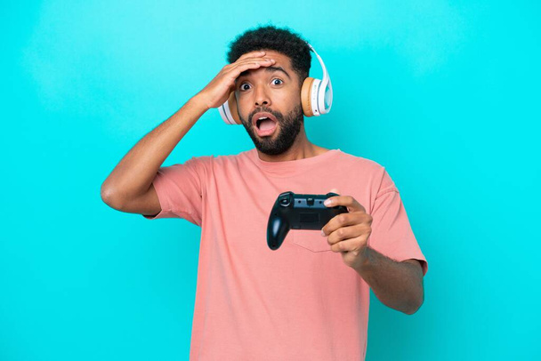 青い背景に隔離されたビデオゲームコントローラーで遊ぶ若いブラジル人の男は側面を見ている間驚きのジェスチャーをしています - 写真・画像