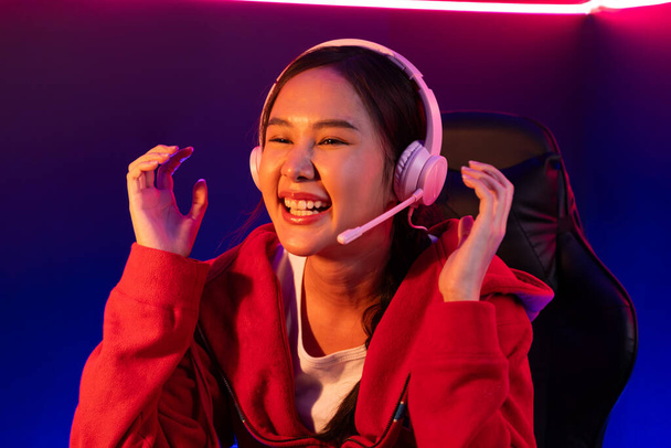 笑顔の美しいアジアの少女ストリーマーのホストチャンネルは,ネオンルームでオンラインゲームと戦うヘッドフォンパステルカラーを身に着けているエスポートの熟練チームプレーヤーに勝利を上げて再生します. ストラテジー. - 写真・画像