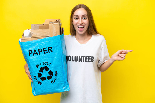Молода біла жінка тримає мішок для переробки, повний паперу, щоб переробляти ізольовані на жовтому тлі, здивована і спрямована пальцем в сторону - Фото, зображення