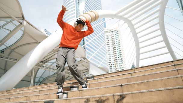Azjatycki hipster w słuchawkach pokazuje kroki podczas skakania po mieście. Profesjonalny tancerz break dance b-boy w mieście. Break dance albo street dance. Sport na świeżym powietrzu 2024. Sprytnie. - Zdjęcie, obraz