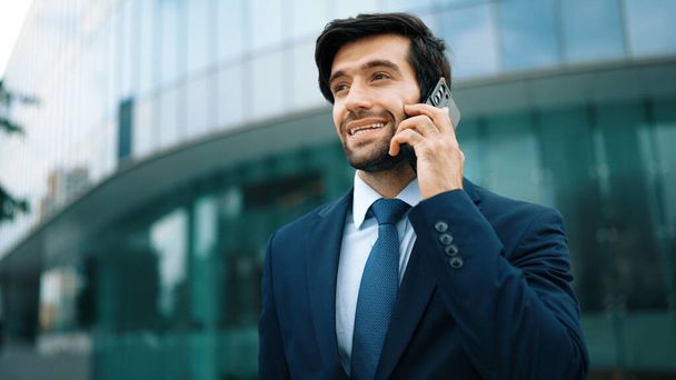 Skilled caucasian project manager calling phone while walking at city with blurred background. Skilled businessman using smart phone talking to project manager with blurred background. Exultant. - Photo, Image