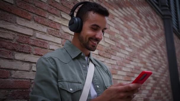 Vista laterale attraente giovane uomo caucasico ascoltare musica con cuffie wireless utilizzando il telefono cellulare appoggiato al muro di mattoni. Millennial maschio guardando felice cellulare sulla strada della città. Copia spazio  - Filmati, video