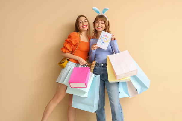 Boldog fiatal nők papír bevásárló táskák és ünnepi képeslap húsvét bézs alapon - Fotó, kép