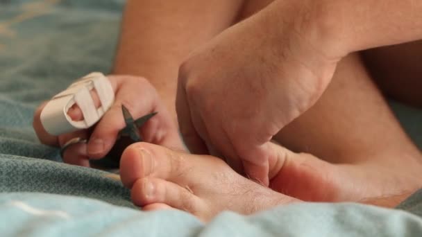 Ένας άντρας κόβει τα νύχια των ποδιών του με σπασμένο δάχτυλο στο κρεβάτι στο σπίτι, κάνοντας πεντικιούρ ο ίδιος - Πλάνα, βίντεο