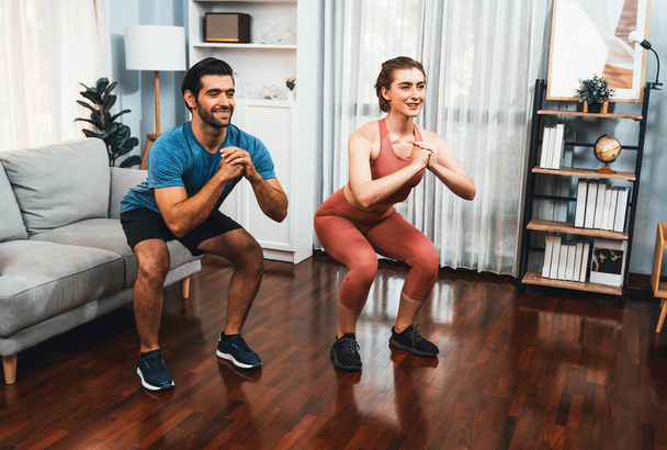 Athletic and sporty couple doing squat together during home body workout exercise session for fit physique and healthy sport lifestyle at home. Gaiety home exercise workout training concept. - Photo, Image