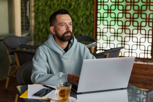 Задумчивый бородатый мужчина в повседневной одежде в кафе с помощью ноутбука. Работает из любой концепции. Онлайн бизнес - Фото, изображение