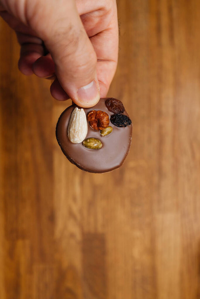 A male hand delicately holds a chocolate-covered cookie adorned with assorted nuts and dried grapes, presenting a delectable treat against a backdrop of fine artistry. - Photo, Image