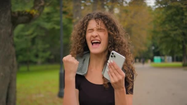 Happy amazed excited surprised woman Caucasian girl hold mobile phone reading news win celebrate victory fortune with smartphone shock yes success happy celebrating after sport outdoors in city park - Footage, Video
