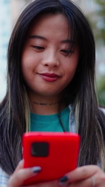 Вертикальна весела азіатська молода жінка сміється, використовуючи мобільний телефон у місті. Покоління z китайської красивої жінки, яка виглядає щасливою на мобільному екрані, насолоджуючись відео в соціальних мережах. Мережеві залежні люди  - Кадри, відео