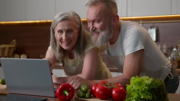 Valkoihoinen hymyilevä pari aikuisia vanha 70s kypsä nainen mies keski-ikäiset puolisot nauravat onnellinen vanhempi perhe käyttää kannettavaa tietokonetta keittiössä tilata tuoreita vihanneksia verkossa supermarket terveellistä ruokaa toimitus kotona - Materiaali, video