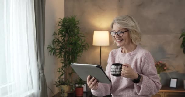 Portret van volwassen blonde blanke vrouw met bril gebruik digitale tablet thuis surfen op internet of bekijk online video vrolijke glimlach slow motion video - Video
