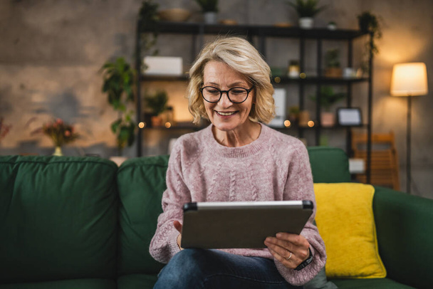 Портрет зрелой белокурой белокурой женщины в очках использовать цифровой планшет на дому просматривать интернет или смотреть онлайн видео счастливая улыбка - Фото, изображение