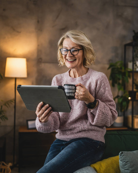 Portrait of mature blonde caucasian woman with eyeglasses use digital tablet at home browse internet or watch online video happy smile - Φωτογραφία, εικόνα