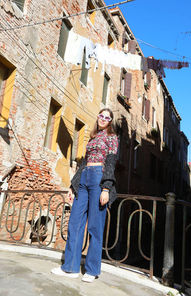 молодая девушка в венецианской маске в синих джинсовых брюках позирует возле домов с одеждой, высушенной на солнце в Италии - Фото, изображение