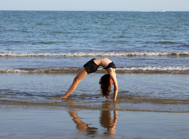 νεαρή κοπέλα εκτελεί γυμναστικές ασκήσεις τοξοβολώντας την πλάτη της και σχηματίζοντας μια καρδιά στην αντανάκλαση στο νερό - Φωτογραφία, εικόνα