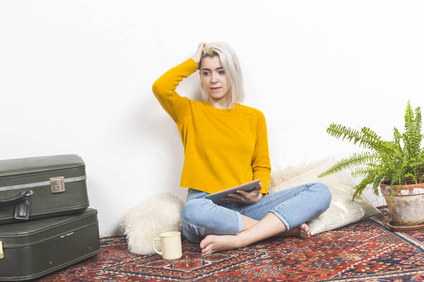 Een casual vrouw met een tablet pauzeert bedachtzaam, haar hand in haar haar, tegen een eenvoudige thuisachtergrond met een vintage koffer en weelderig groen, weerspiegelt gemak en reflectie. - Foto, afbeelding