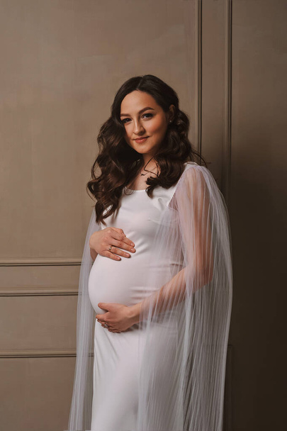 Szczęśliwa kobieta w ciąży w pięknej białej sukience stojącej w pobliżu ściany w photostudio i trzymając jej brzucha w ciąży z rękami. Koncepcja szczęśliwego macierzyństwa i rodzicielstwa - Zdjęcie, obraz