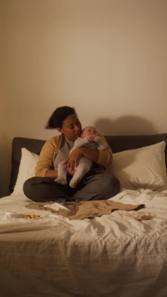 Full vertical shot of happy young African American mother in cardie and pyjamas sitting on bed at home, holding up baby boy in bodysuit, cooing and kissing him - Footage, Video