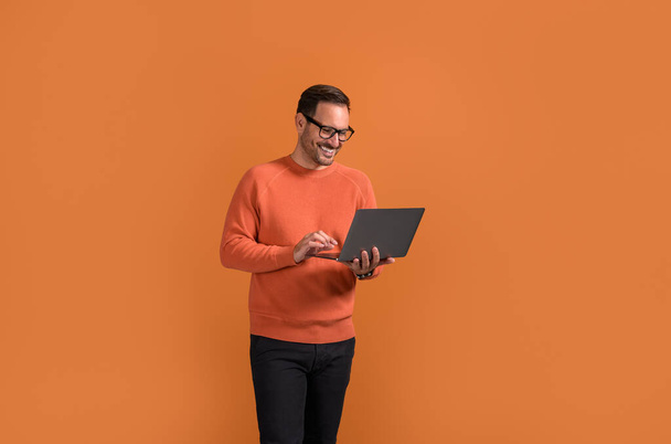 Gerente masculino sonriente revisando correos electrónicos sobre el ordenador portátil mientras está de pie con confianza en el fondo naranja - Foto, Imagen