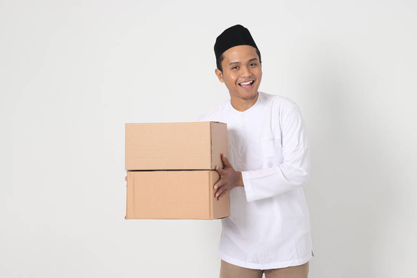 Portrait of excited Asian muslim man in koko shirt with peci carrying cardboard box. Going home for Eid Mubarak. Isolated image on white background - Photo, Image
