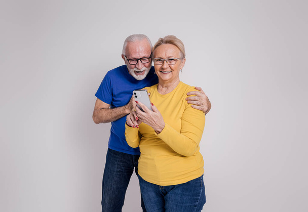 Ενθουσιασμένος ηλικιωμένος σύζυγος που διαβάζει καλά νέα από το έξυπνο τηλέφωνο που κρατά η χαμογελαστή σύζυγος σε λευκό φόντο - Φωτογραφία, εικόνα
