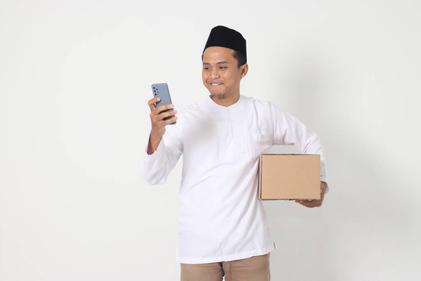 Портрет збудженого азіатського мусульманського чоловіка в сорочці з пивом, що несе картонну коробку, тримаючи мобільний телефон. Повертаюся додому до Ід Мубарака. Ізольоване зображення на білому тлі - Фото, зображення