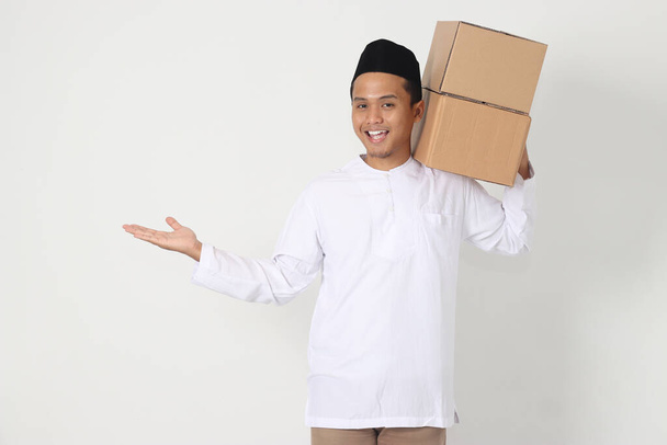 Retrato de hombre musulmán asiático emocionado en camisa koko con peci que lleva caja de cartón mientras señala hacia el lado. Voy a casa por Eid Mubarak. Imagen aislada sobre fondo blanco - Foto, imagen