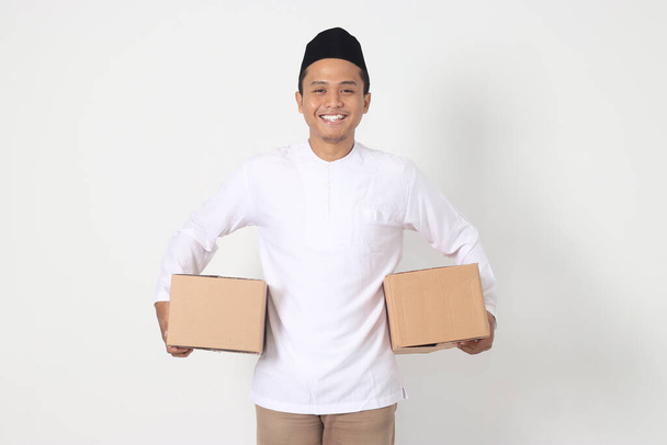 Portrait of attractive Asian muslim man in koko shirt with peci feeling grateful and peacful while carrying cardboard box. Going home for Eid Mubarak. Isolated image on white background - Photo, Image
