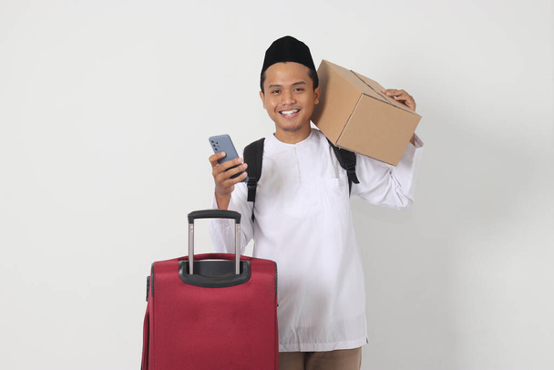Ritratto di uomo musulmano asiatico eccitato che trasporta scatola di cartone e valigia mentre tiene lo smartphone. Vado a casa per Eid Mubarak. Immagine isolata su sfondo bianco - Foto, immagini