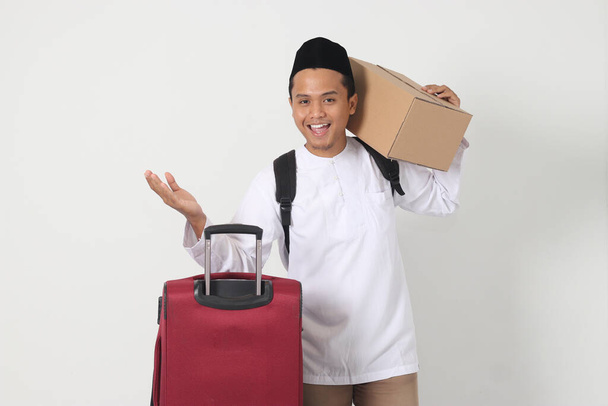 Ritratto di uomo musulmano asiatico eccitato che porta scatola di cartone e valigia mentre punta lateralmente con il dito. Vado a casa per Eid Mubarak. Immagine isolata su sfondo bianco - Foto, immagini
