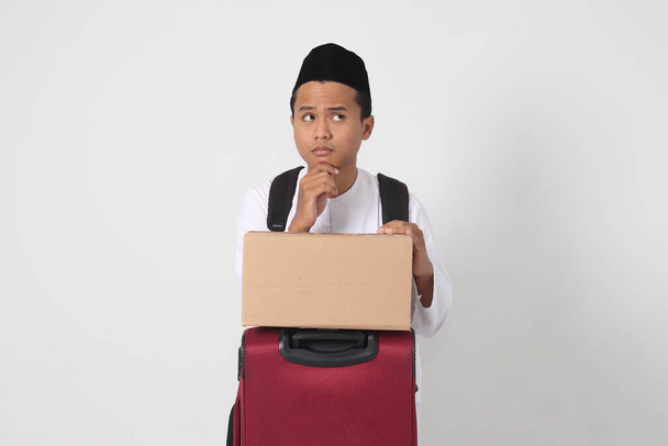 Ritratto di uomo musulmano asiatico confuso che tiene valigia e scatola di cartone e pensa con mano sul mento. Vado a casa per Eid Mubarak. Immagine isolata su sfondo bianco - Foto, immagini