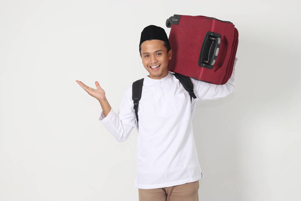 Retrato de un hombre musulmán asiático emocionado llevando una maleta apuntando con la mano hacia un lado. Voy a casa por Eid Mubarak. Imagen aislada sobre fondo blanco - Foto, imagen