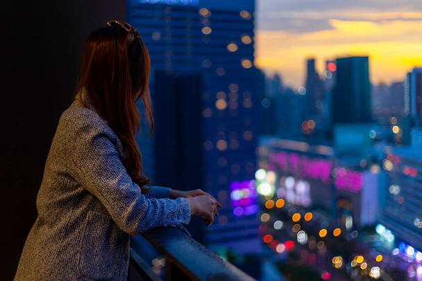 Mujer asiática joven disfrutando de la vista de la noche desde su balcón mientras mira el paisaje urbano de rascacielos por la noche para la vivienda y el estilo de vida moderno - Foto, imagen