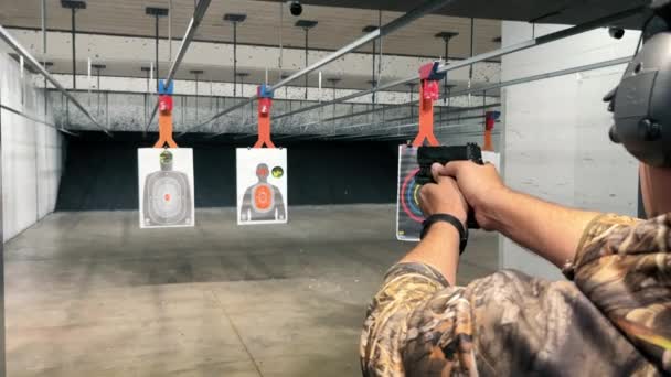 Hombre practicando en el campo de tiro interior, un hombre en traje de camuflaje apuntando con una pistola a objetivos en un campo de tiro interior - Imágenes, Vídeo