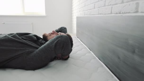 Homem descansando pacificamente na cama, um homem barbudo se deita com os braços cruzados atrás da cabeça em uma cama em um quarto brilhante, olhos fechados em repouso. - Filmagem, Vídeo