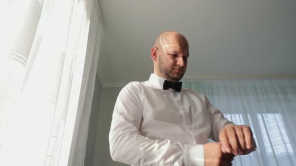 Gentleman Adjusting Watch Before Event, un hombre reflexivo con una camisa blanca ajusta su corbata de lazo, bañado en suave luz solar por la ventana - Metraje, vídeo