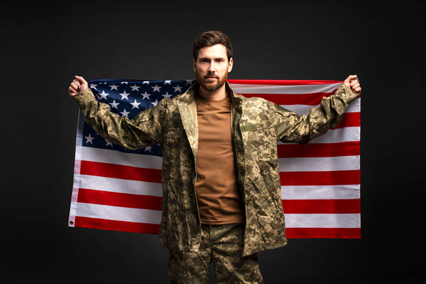 Портрет серйозної людини, впевнений солдат у військовій камуфляжній формі, що тримає американський прапор ізольовано на чорному тлі. Концепція Дня виборів США  - Фото, зображення