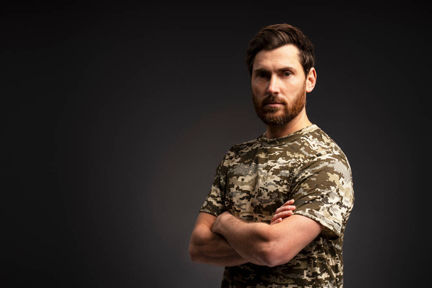 Handsome bearded man, serious soldier holding arms crossed wearing military camouflage uniform looking at camera isolated on black background, copy space  - Photo, Image