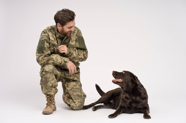 Un homme souriant barbu attrayant portant un uniforme de camouflage militaire et un chien du Labrador isolé sur fond blanc. Amour, amitié, concept de dressage de chiens  - Photo, image