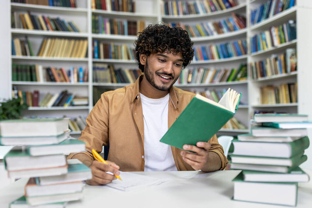 A cheerful graduate student with curly hair is immersed in exam preparation, surrounded by stacks of books at a campus library. He takes notes and reads intently, conveying a sense of learning and - 写真・画像