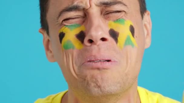 Primer plano de vídeo en estudio con croma de un hombre muy nervioso con una bandera jamaicana pintada en la cara, apoyando equipo jamaicano durante un partido difícil que al final se pierde - Metraje, vídeo